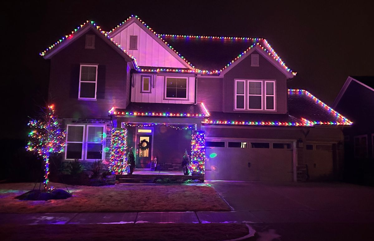 Two-story home outlined in multicolor C9 bulbs with wrapped porch columns and a lit tree